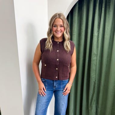 Woman wearing a sleeveless brown top and blue jeans standing in front of a green curtain.