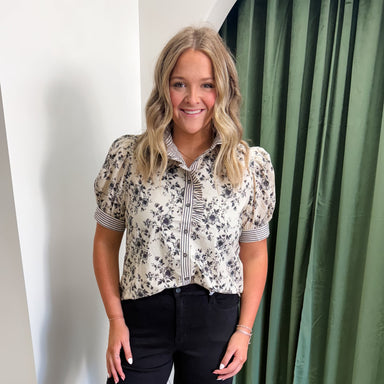 Woman wearing a floral blouse and black pants with white boots in front of green curtains.